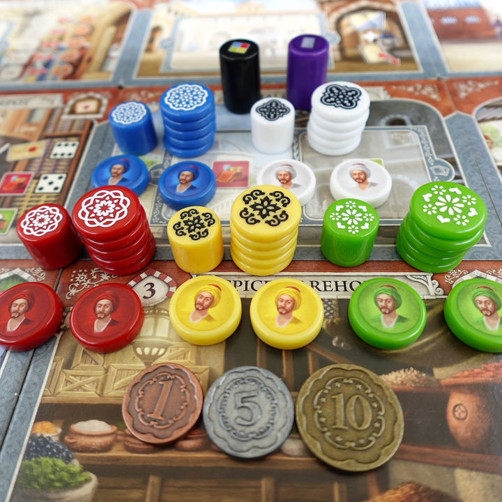 A photo of the GeekUp upgraded bits for use with the board game Istanbul. The pieces are arranged in stacks by color, with a single black and purple token in the background. Each ground has a stack of engraved disks with a Turkish tile design, and a token with an illustration of a man in a turban. Three metal coins are placed in the front, each a different size, color, and value.