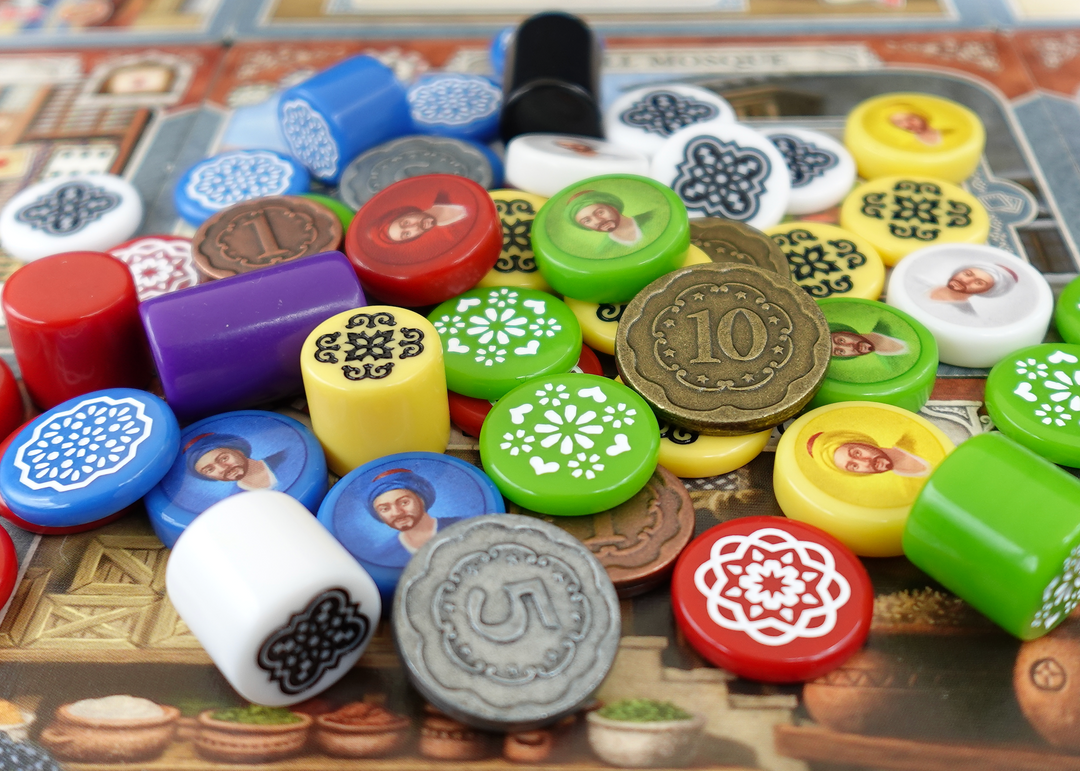 An overhead photo of a jumble of GeekUp bits for use with the board game Istanbul. Some tokens have an illustration of a man in a turban printed on them. Some tokens are engraved and filled with white paint and some are metal coins. All of the tokens are sitting on top of tiles that create the game's main board.