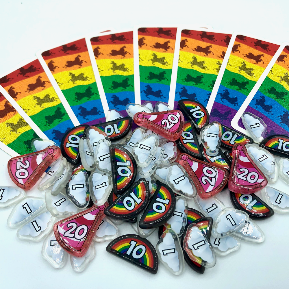 A photo of upgrades for the card game L.A.M.A., featuring a pile of plastic tokens in the shapes of rainbows, clouds, and party hats. Behind is an array of cards from the game with rainbow backs, and everything photographed on a white background.