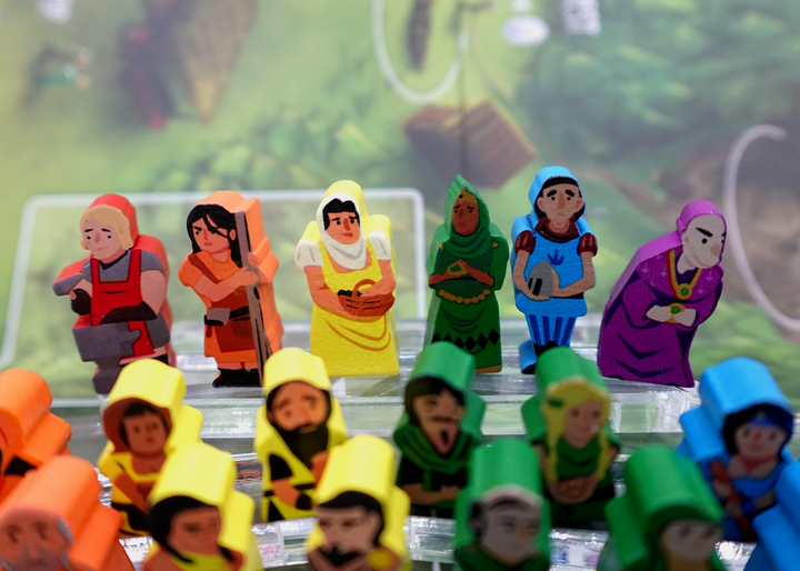 A close up photo of a group of people tokens made from wood, for use with the board game Architects of the West Kingdom. These tokens are painted different colors, and each has its own shape, clothes, hair, and expressions painted on one side.