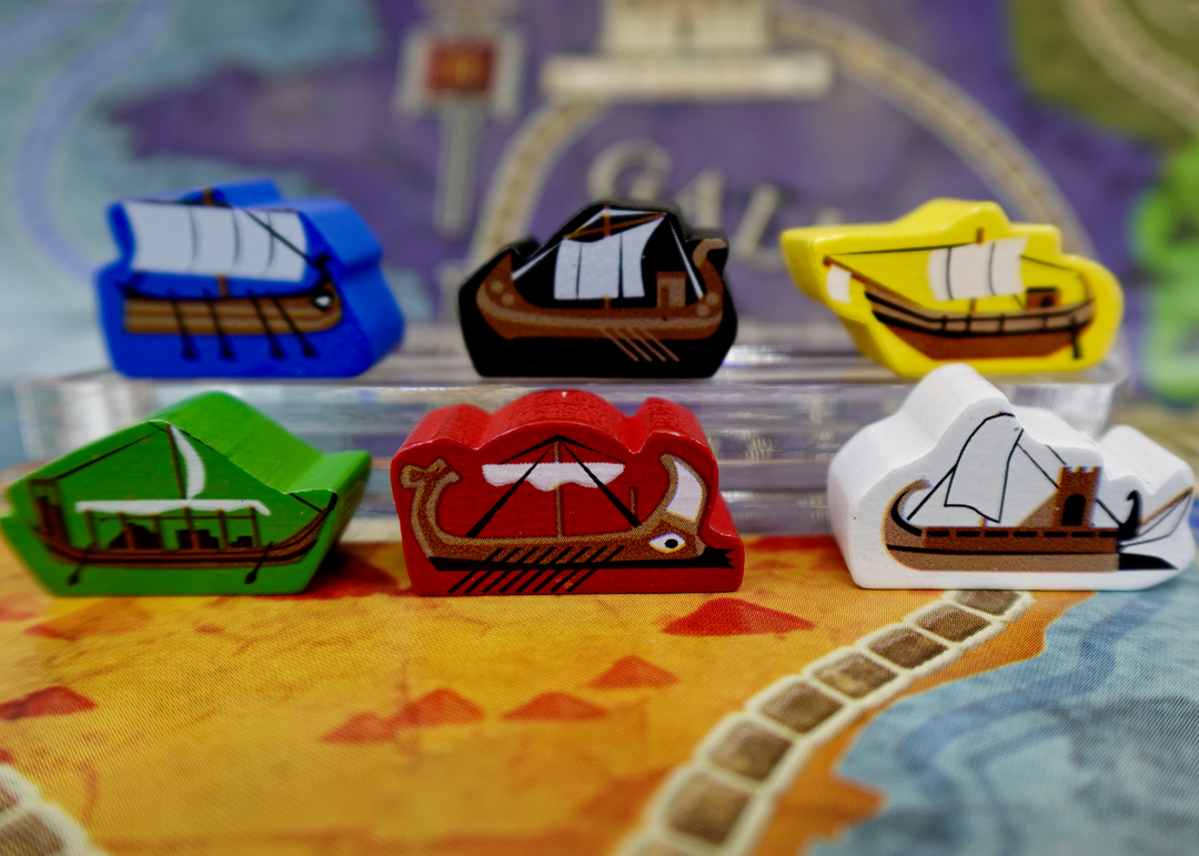 A pile of wooden tokens for use with the board game Concordia, featuring boats from ancient Greece, painted in a variety of background colors.