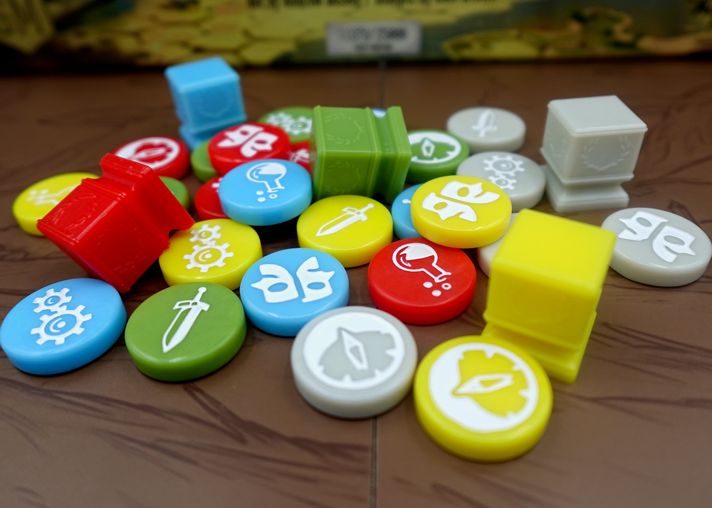 A side-view photo of a jumble of GeekUp bits for use with the board game Tapestry. Most bits are circular disks in a variety of colors, each with an engraved symbol in the center. The rest of the tokens are square, plastic columns with a laurel wreath printed on each side.