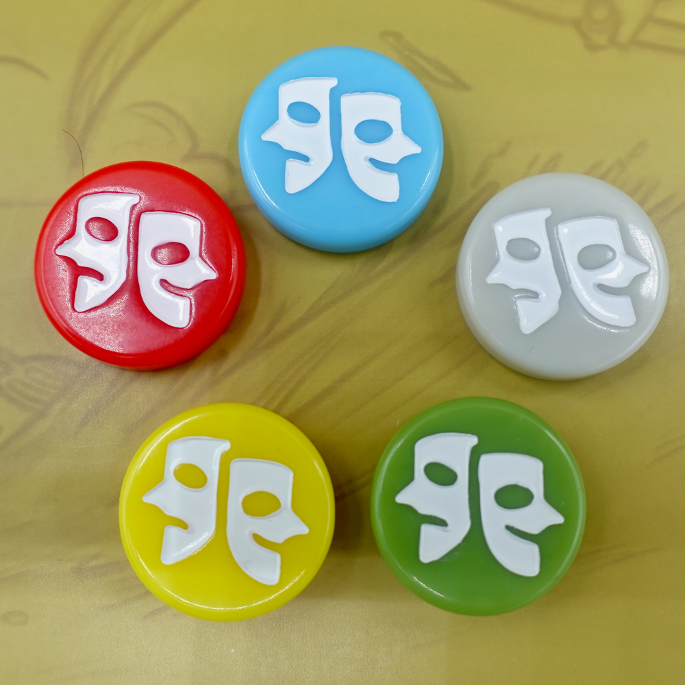 A photo of five, plastic tokens, each with an engraving of drama masks, sitting on a cardboard background with a tan illustration of wheat and grass.