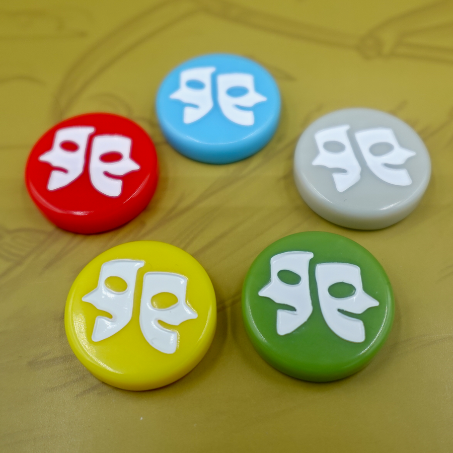 A photo of five, plastic tokens, each with an engraving of drama masks, sitting on a cardboard background with a tan illustration of wheat and grass.