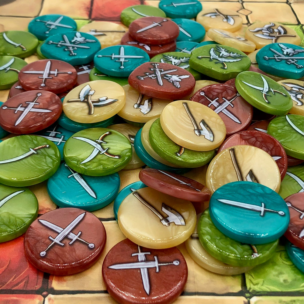A photo of plastic tokens for use with the board game Tash-Kalar in a variety of colors, all with weapons of different types printed on the tops and bottoms, sitting on a game board printed with a square grid.