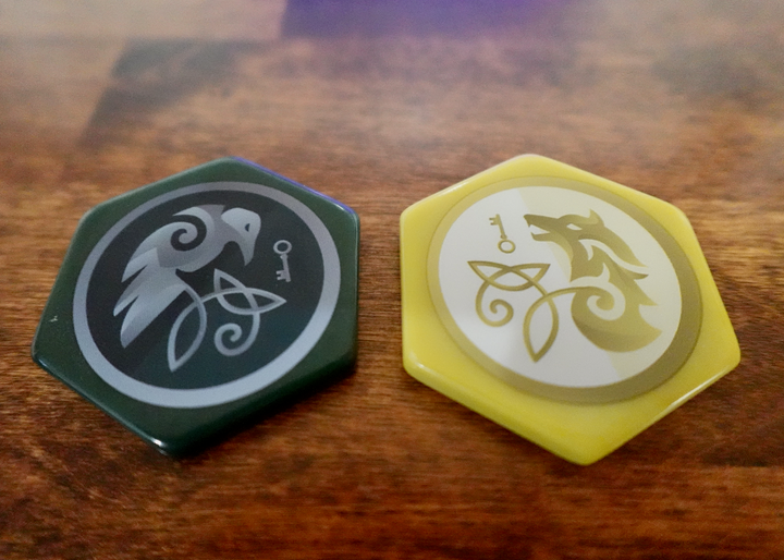 A closeup photo of two upgraded, hexagon-shaped tokens for use with the board game War Chest. One is dark green with an illustration of a stylized raven and key. The other is dark gold with a stylized illustration of a wolf and a key.