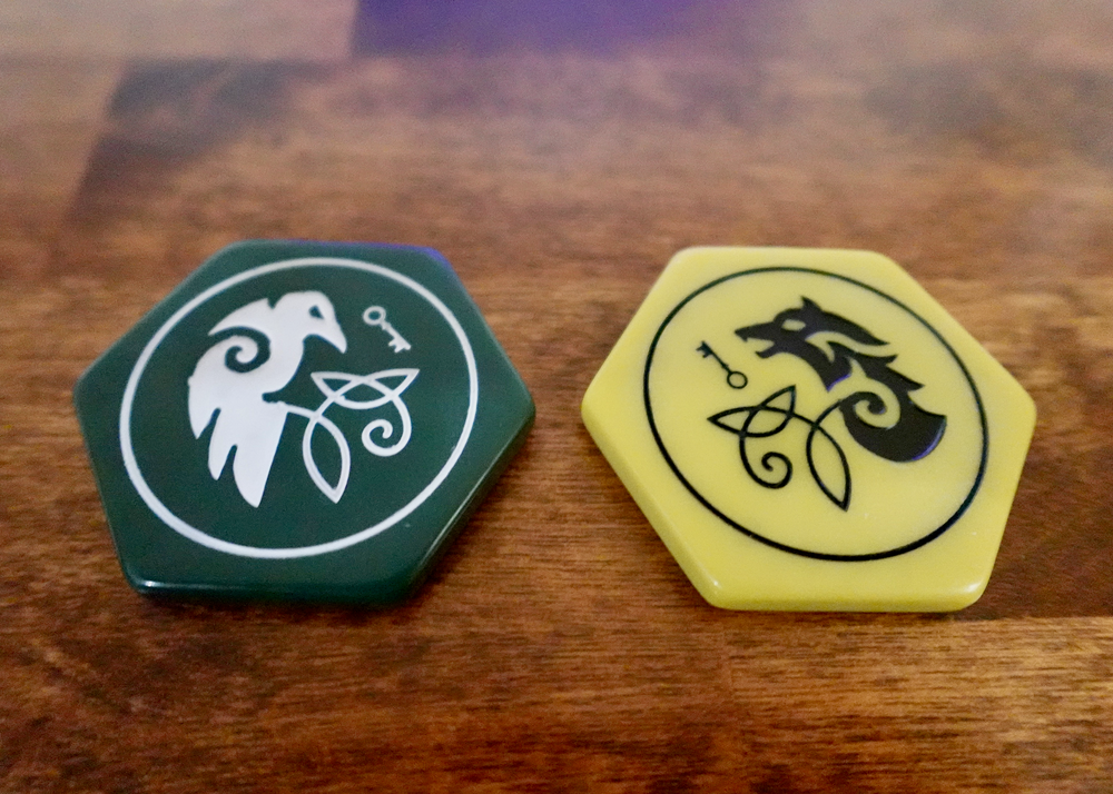 A closeup photo of two upgraded, hexagon-shaped tokens for use with the board game War Chest. One is dark green with a engraving in white paint of a stylized raven and key. The other is dark gold with a stylized engraving in black paint of a wolf and a key.
