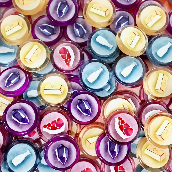 An overhead photo of a jumble of GeekUp tokens for use with the board game Wyrmspan. Each token is made from transparent, colored plastic, and has an illustration of gold, milk, meat, or a crystal printed in the center.