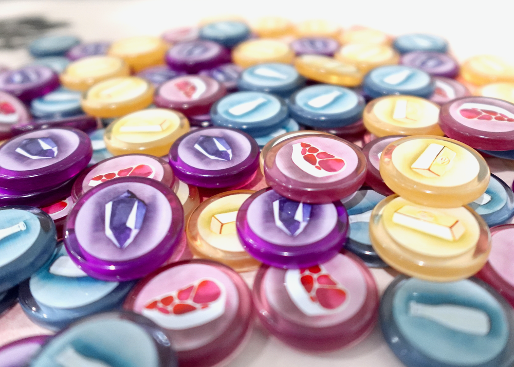 A side-view photo of a jumble of GeekUp tokens for use with the board game Wyrmspan. Each token is made from transparent, colored plastic, and has an illustration of gold, milk, meat, or a crystal printed in the center.