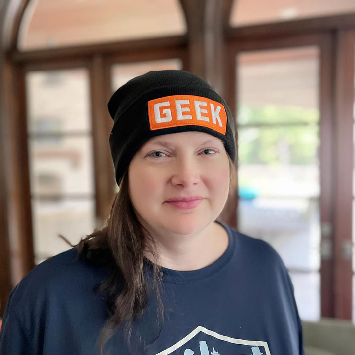 A photo of a Caucasian woman standing in front of windows, wearing a black knit ski cap decorated with an orange and white patch that reads GEEK.
