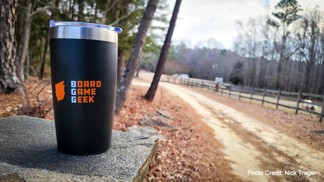 A black and silver travel mug, with a clear, push top above the silver rim, sitting outside on a stone wall with a forest and dirt road in the background. The mug is decorated with an orange logo and the words "BoardGameGeek".