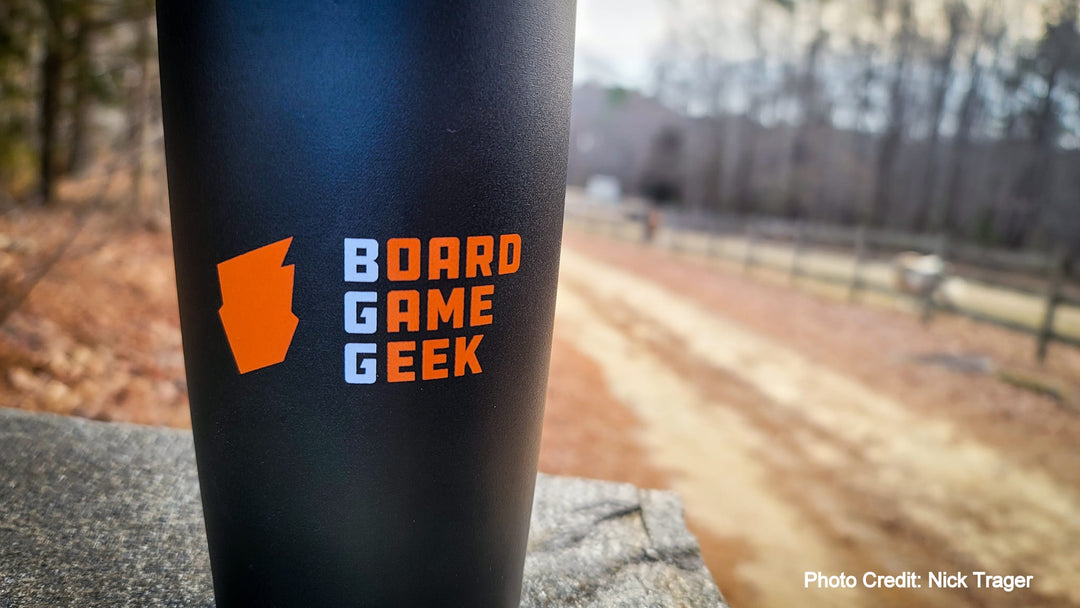 A closeup of the side of a black travel mug, sitting outside on a stone wall with a forest and dirt road in the background. The mug is decorated with an orange logo and the words "BoardGameGeek".