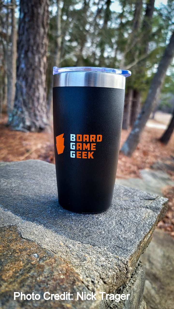 A black and silver travel mug, with a clear, push top above the silver rim, sitting outside on a stone wall with a forest in the background. The mug is decorated with an orange logo and the words "BoardGameGeek".