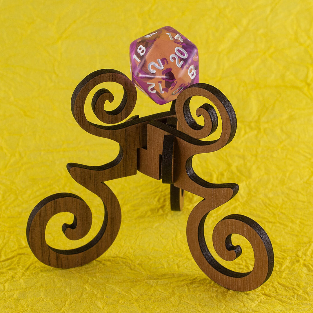 A laser-cut wooden, tripod stand with decorative curves, holding up a 20-sided die, on a yellow background