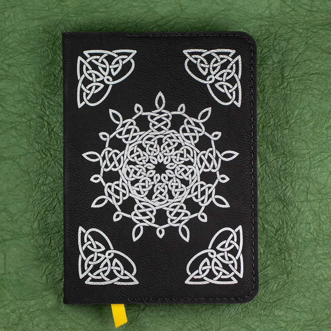 A black leather journal with a white Celtic pattern, a yellow ribbon bookmark, sitting on a green background..