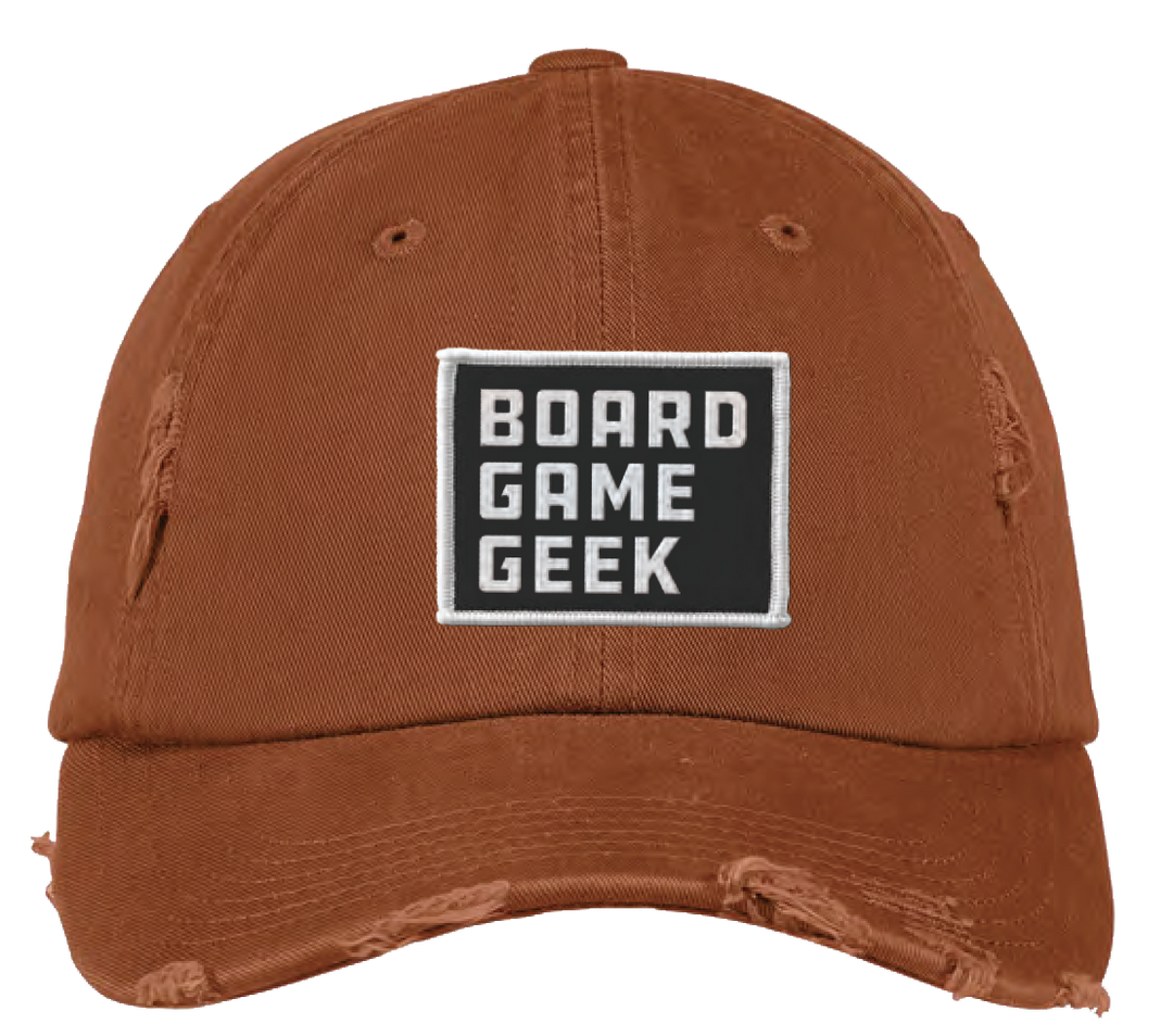 A baseball cap that is burnt orange with intentional distressing, and a black-and-white patch that says "BoardGameGeek" in the front center.