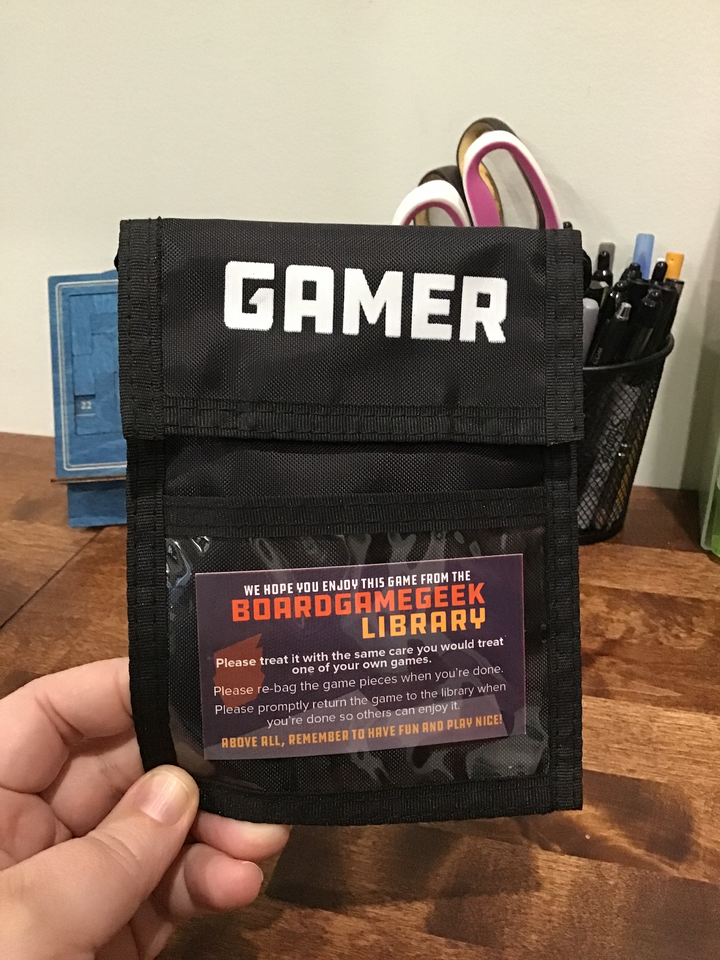 A photo of a black neck wallet with black trim. The top flap is closed down and printed with a logo and the word "GAMER" at the top. A business card for the "BoardGameGeek Library" sits in the middle of a plastic window pocket at the bottom.