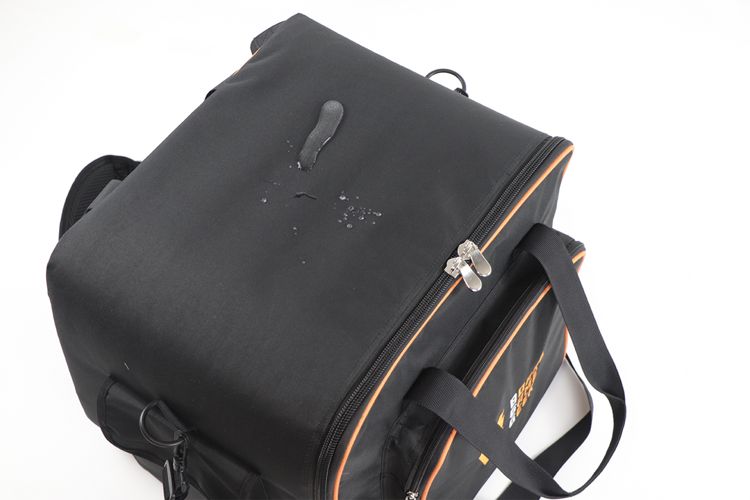 A side view of a black carry bag and backpack with orange trim, as well as a splatter of water that is beaded on top of the fabric.