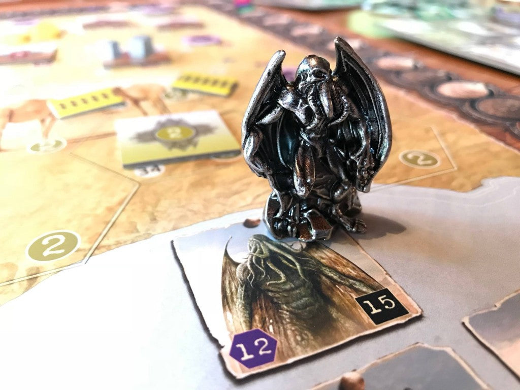 A close-up photo of the metal upgrade token of Cthulhu for the board game AuZtralia. The figurine is a standing figure with wings and tentacles draped from its head, placed next to the cardboard token from the game with the same image. 