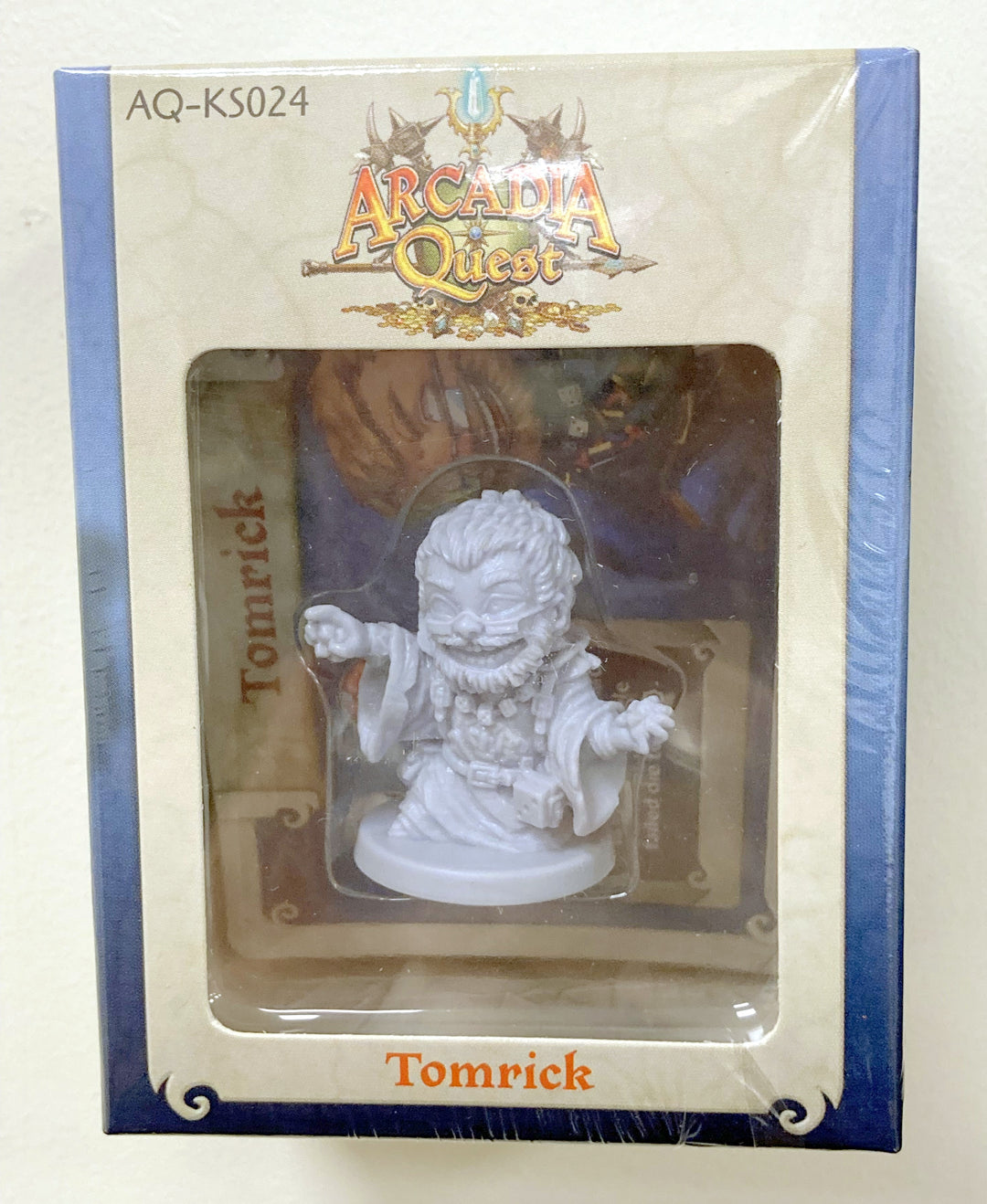 A picture of a miniature figurine in its box for use with the board game Arcadia Quest. This figure is labelled Tomrick, and features a bearded man in long robes.