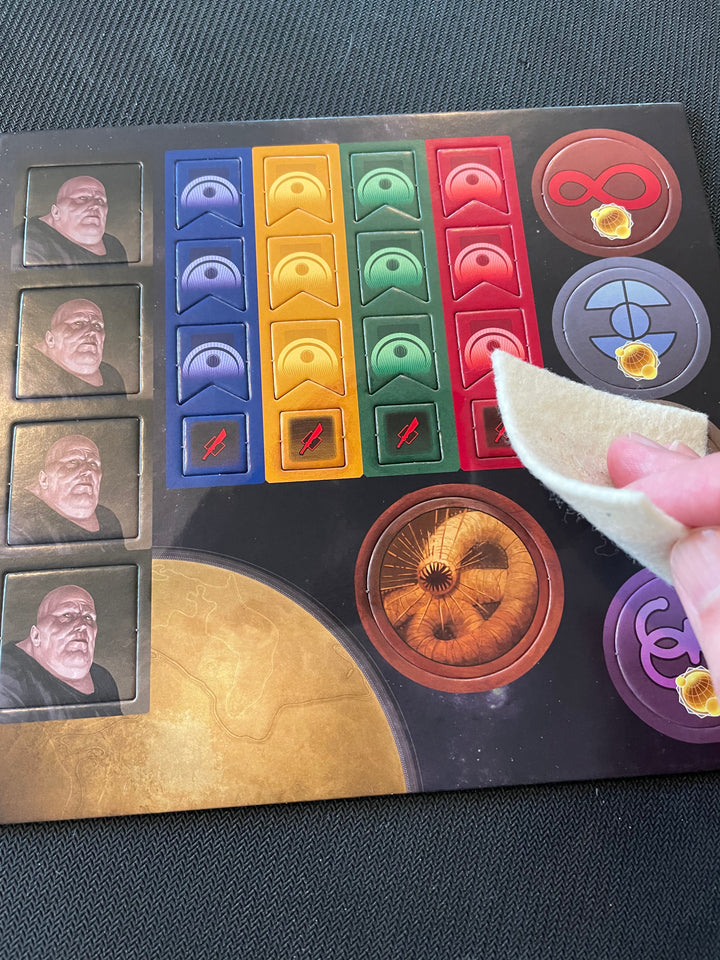 A photo of a punchboard from a board game, and a hand with a felt square rubbing the surface.