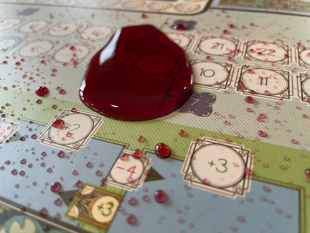 A close up photo of a game board treated with BGShield, with a pool and multiple small splattered of red wine sitting in beads on top.