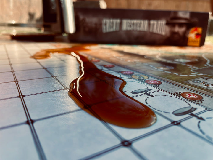 A close up photo of a puddle of coffee sitting on the game board for the board game Great Western Trail, with the game box visible in the background.