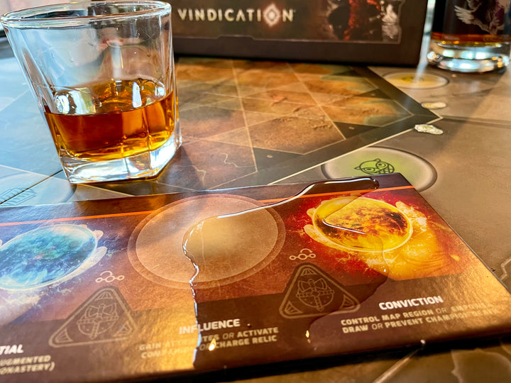 A photo of a puddle of liquid on top of a player board and game board for the board game Vindication, next to a tumbler of liquid and the game box in the background.