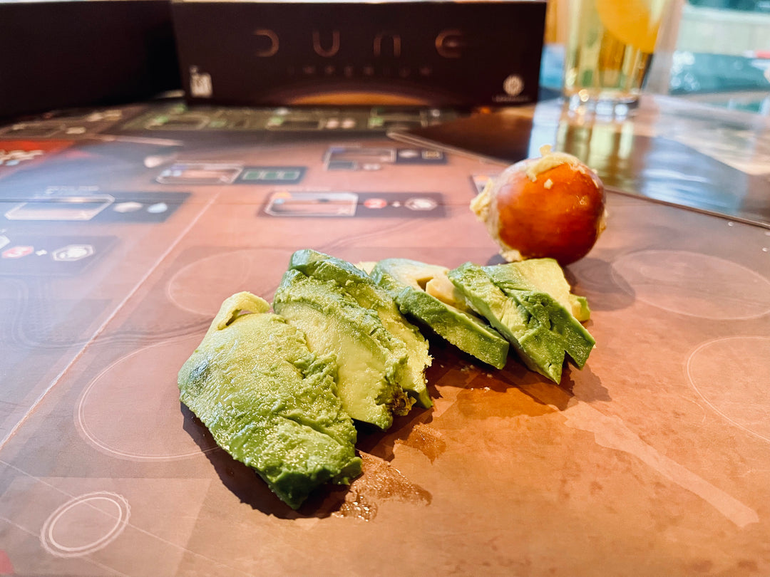 A photo of pieces of a sliced avocado and an avocado pit sitting directly on the board for the board game Dune Imperium, with the game box visible in the background.