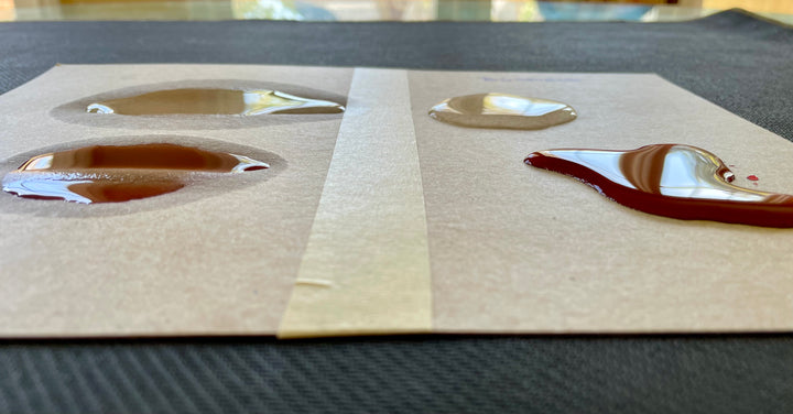 A side angle photo of a side-by-side comparison of two cardboard surfaces with two liquids on top: one clear and one red. The left sample shows the liquids sinking in and getting absorbed into the cardboard. The right side is labeled "BGShield" and shows the two liquids suspended on the top of the cardboard.
