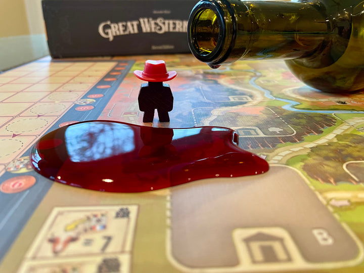 A close up photo of a puddle of red wine sitting on the game board for the board game Great Western Trail, next two a figure from the game and a wine bottle, with the game box visible in the background.