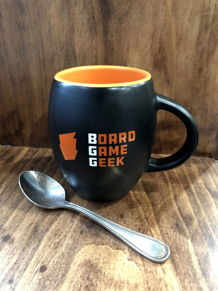 A barrel-shaped, matte black coffee mug, with orange on the inside, labeled with "BoardGameGeek" on the outside, and against a polished wooden background with a teaspoon laying in front.