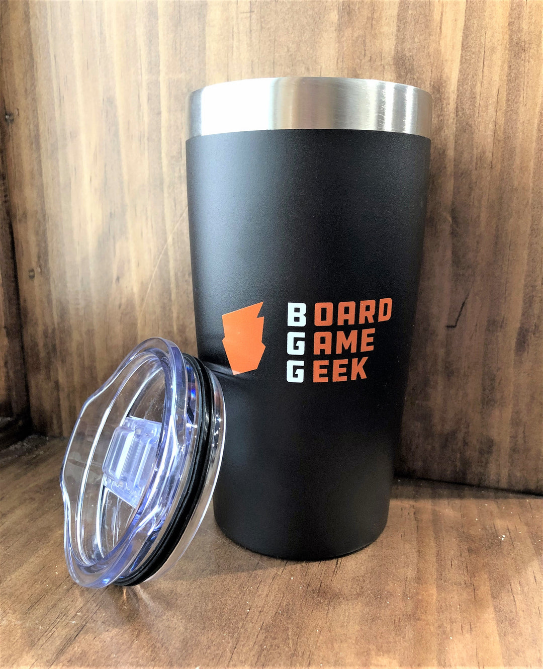 A black and silver travel mug sitting on a wooden background, with a clear, push top resting on the side. The mug is decorated with an orange logo and the words "BoardGameGeek".