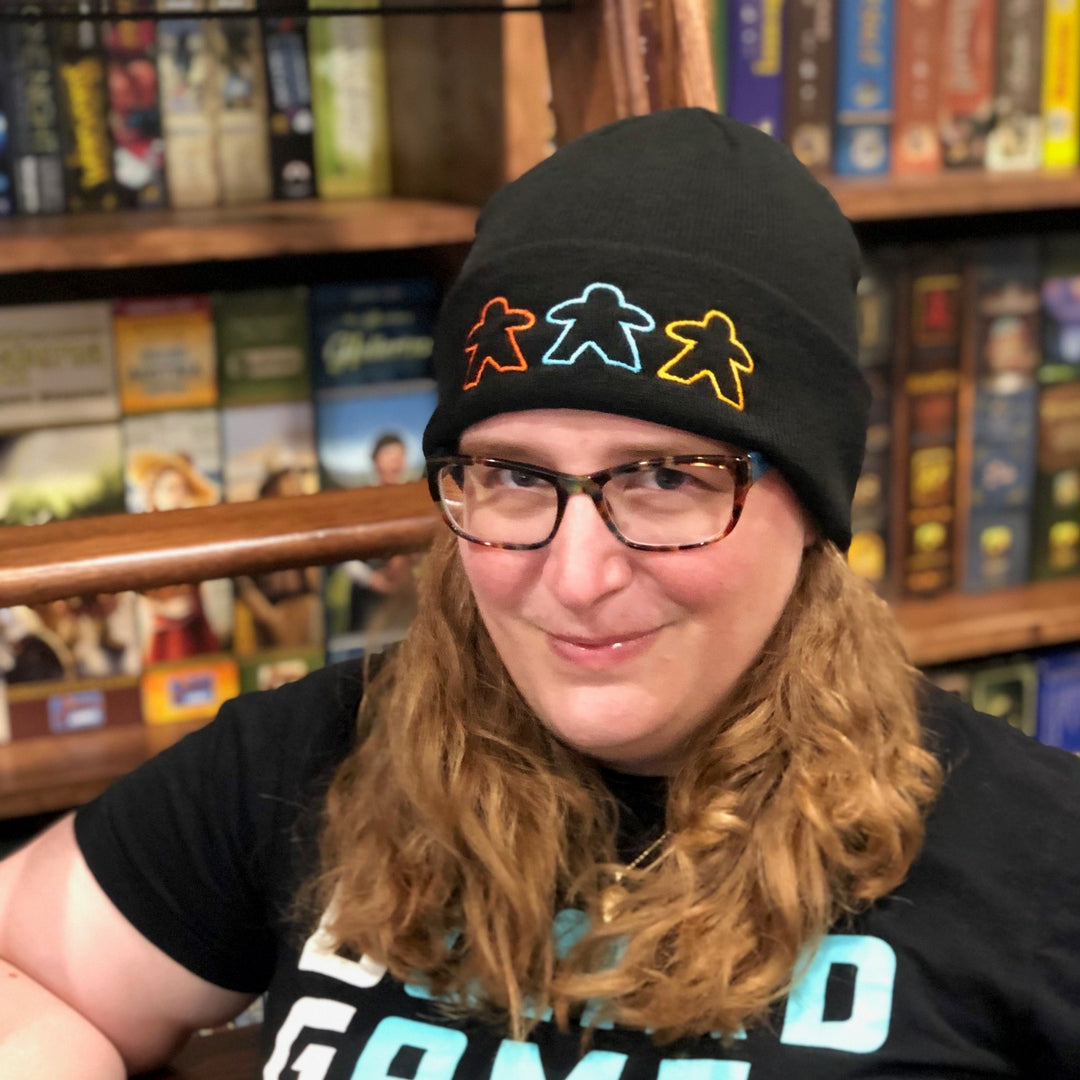 A photo of a caucasian woman wearing a black knit ski cap decorated with three, outlined meeples in various colors, standing in front of a bookcase of board games.