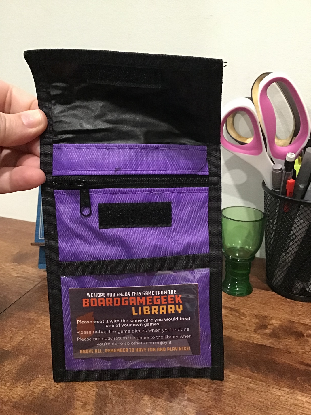 A photo of a purple neck wallet with black trim, with the top flap flipped up, multiple pockets, and a business card for the "BoardGameGeek Library" in a plastic window pocket at the bottom.