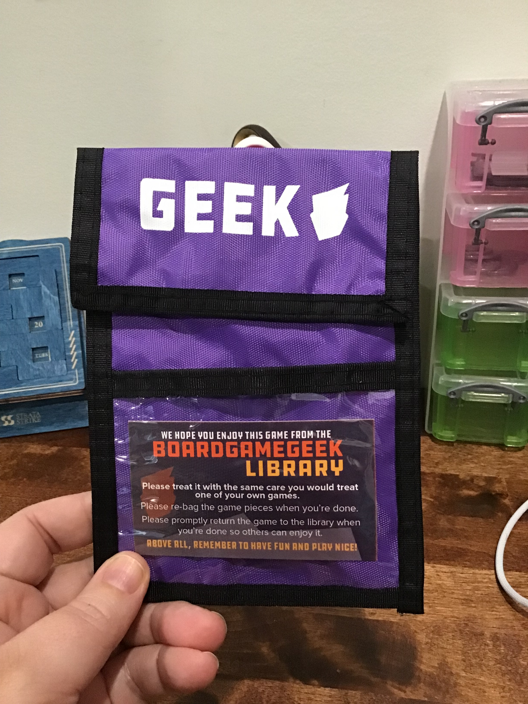 A photo of a purple neck wallet with black trim. The top flap is closed down and printed with a logo and the word "GEEK" at the top. A business card for the "BoardGameGeek Library" sits in the middle of a plastic window pocket at the bottom.