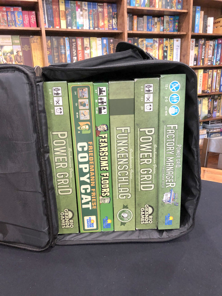 A black carry bag and backpack, sitting on a padded tabletop, with six board games stacked horizontally inside, and multiple bookcases of board games in the background.