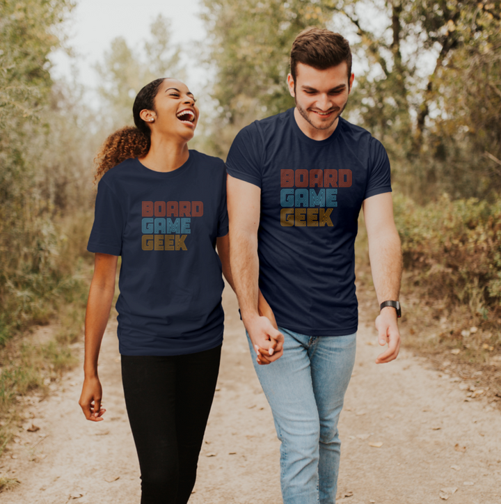 A photo of a woman and man outside, holding hands and smiling, both wearing the same t-shirt. The shirt is dark navy, and has the words "BoardGameGeek" written in three different colors on the front.